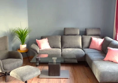 Gemütlicher Wohnzimmer einer Ferienunterkunft in Bolsdorf (bei Hillesheim) mit dunkelgrauem Sofa, Relaxsessel und Fenster mit Vorhängen.